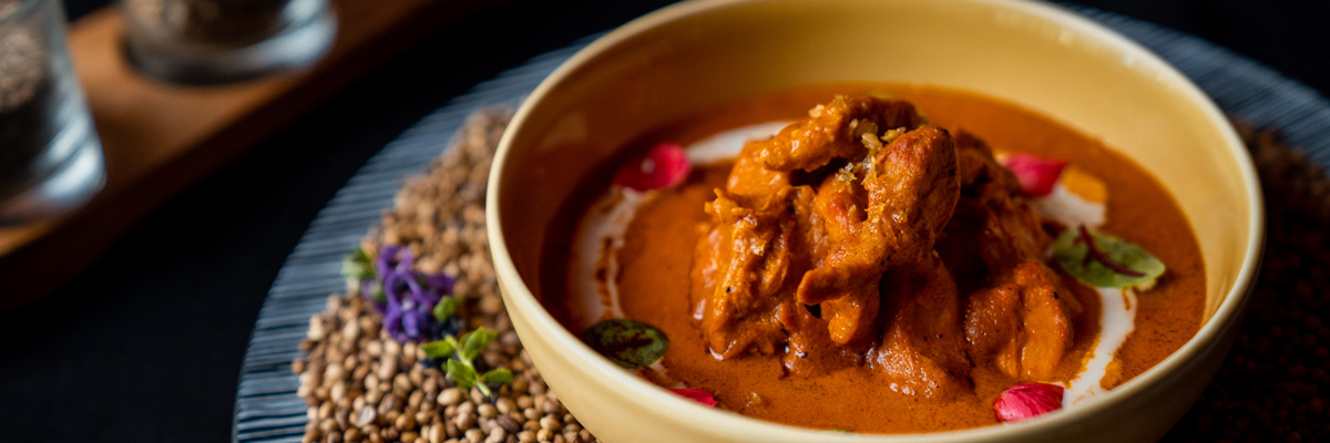 Traditional Butter chicken in a yellow bowl on top of coriander bed on a glass striped plate with black background at Queen’s Tandoor Seminyak